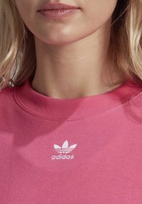 Růžová bavlněná mikina s žebrovaným kulatým výstřihem, na prsou s bílým logem Adidas a motivem listu. Je vidět měkká textura látky.