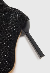 Chaussure à talon aiguille noire avec une surface texturée ornée de petits strass noirs, un bout pointu élégant et un talon aiguille effilé.