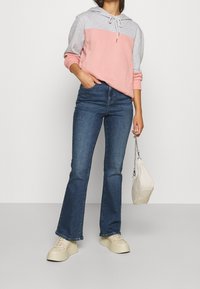 Person som bär en rosa och grå hoodie, blå utställda jeans, beige plattformssneakers och håller en liten vit handväska med kedjesnodd.