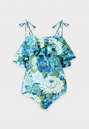 Kvinde stående barfodet iført en blå blomsterprint badedragt med flæse-top og justerbare skulderstropper mod en hvid baggrund.