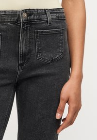 Gros plan sur la taille et la main d'une personne portant un jean en denim noir taille haute avec un bouton à l'avant et une poche.