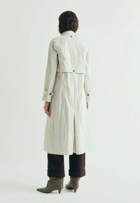 Person står med ryggen mod, iført en lang cremefarvet trenchcoat, mørke bukser med opkropper og beige, spidsede ankelstøvler på hvid baggrund.