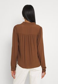 Femme aux longs cheveux noirs portant un chemisier marron à manches longues et un pantalon blanc, debout de dos face à la caméra sur un fond blanc.