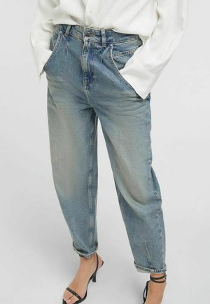 Jeans azzurri chiari a vita alta con vestibilità comoda e risvoltini, indossati con una camicia bianca e sandali neri con tacco e cinturini. Mani nelle tasche anteriori.