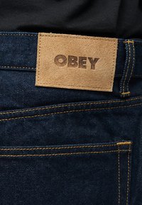 Καφέ σουέντ πάτς με την ανάγλυφη λέξη "OBEY" σε σκούρο μπλε τζιν με πορτοκαλί ραφές, φορεμένο με μαύρο ρούχο πάνω από τη μέση.