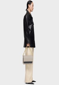 Mujer con chaqueta de cuero negra y pantalones beige, sosteniendo un bolso beige con una cadena dorada, de perfil frente a un fondo liso.