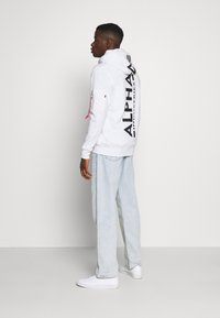 Sweatshirt à capuche blanc avec le texte noir "ALPHA" au dos, associé à un jean en denim bleu clair et des baskets blanches. Comprend une poche zippée sur la manche.