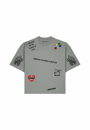 Frau trägt übergroßes graues T-Shirt mit aufgedruckten Phrasen, Emojis, brennenden Würfeln und Herzgrafiken, kombiniert mit beigen Hosen und gelber Sonnenbrille.