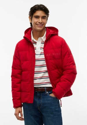 Joven sonriente con chaqueta acolchada roja con capucha, camiseta polo blanca a rayas, vaqueros azules y cinturón marrón con la mano en el bolsillo.