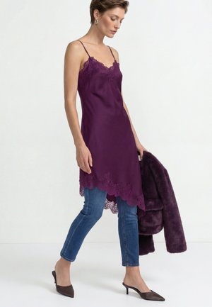 Donna che indossa un vestito scivolato viola con bordo in pizzo sopra jeans blu, tiene in mano un cappotto di pelliccia viola abbinato, camminando con mules con tacco a punta.