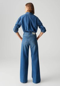Chemise en denim bleu foncé, coupe oversize avec des manches retroussées. Jeans taille haute à jambes larges en bleu moyen avec deux poches arrière et une marque minimale.