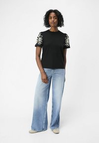 Svart t-shirt med blommigt vitt broderi på ärmarna, kombinerad med ljusblå vida jeans och beiga skor. Slät textur, avslappnad design.