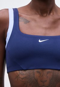 Κοντινό πλάνο ενός ατόμου που φοράει ένα σκούρο μπλε αθλητικό σουτιέν Nike με λευκές τιράντες, που δείχνει ένα τατουάζ λιονταριού στην άνω κοιλιακή χώρα και ένα μερικώς ορατό τατουάζ στον ώμο.