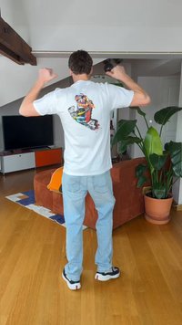 Man wijst met duimen naar een kleurrijke afbeelding op de achterkant van een wit T-shirt in een woonkamer met planten en een tv.