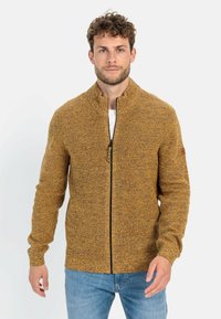 Gelb-brauner, gestrickter Reißverschluss-Pullover mit hohem Kragen, gerippten Bündchen und strukturiertem Finish, getragen über einem weißen Hemd und blauen Jeans.