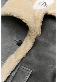 Abrigo de cuero gris con un cuello de borrego marrón claro, que presenta un botón negro y una solapa de bolsillo, mostrando una textura suave y colores contrastantes.