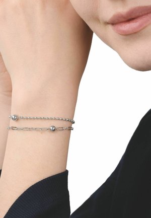 Deux bracelets en chaîne argentée avec des perles rondes sur un poignet, à côté d'une partie du visage souriant d'une personne portant une manche noire.