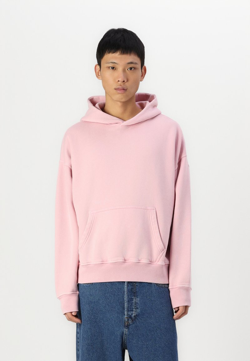 Sweatshirt à capuche rose clair en tissu doux. Présente une poche avant et des poignets côtelés. Associé à un short en denim ample.