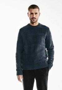 Pull en maille bleu marine avec des rayures horizontales texturées, un col et des poignets côtelés, et une coupe décontractée. Porté sur un pantalon sombre.