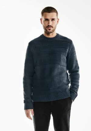 Suéter de punto azul marino con rayas horizontales texturizadas, cuello y puños acanalados, y un ajuste holgado. Llevado sobre pantalones oscuros.