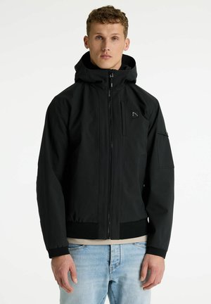 Fiatal férfi fekete cipzáras kapucnis dzsekit visel elülső és ujjrészen zsebekkel, világoskék farmerrel párosítva, fehér háttér előtt áll.