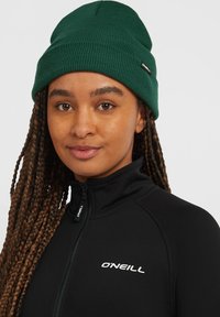Groene geribbelde beanie met een omslag, gedragen over een zwarte jack met ritssluiting met een wit logo. Close-up van de details van de hoed en het jack.