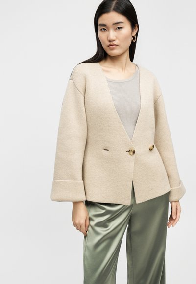 Γυναίκα που φοράει μπεζ ζακέτα με κουμπιά, ανοιχτό γκρι ριγωτό τοπ, χακί σατινέ παντελόνι και χρυσούς κρίκους σκουλαρίκια μπροστά σε ένα μονόχρωμο φόντο.