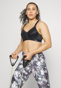 Svart sports-bh med mesh-detaljer, kombinert med mønstrede leggings i nyanser av grått, lilla og hvitt. Motstandsbånd holdt i hendene.