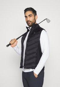 Chaleco acolchado negro con textura de rombos, cierre de cremallera y bolsillos laterales; llevado sobre una camisa blanca de manga larga que presenta un palo de golf.