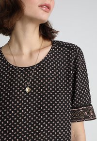 Vrouw die een zwart patroonjurk met korte mouwen draagt en een gouden ketting met een rond hangertje.