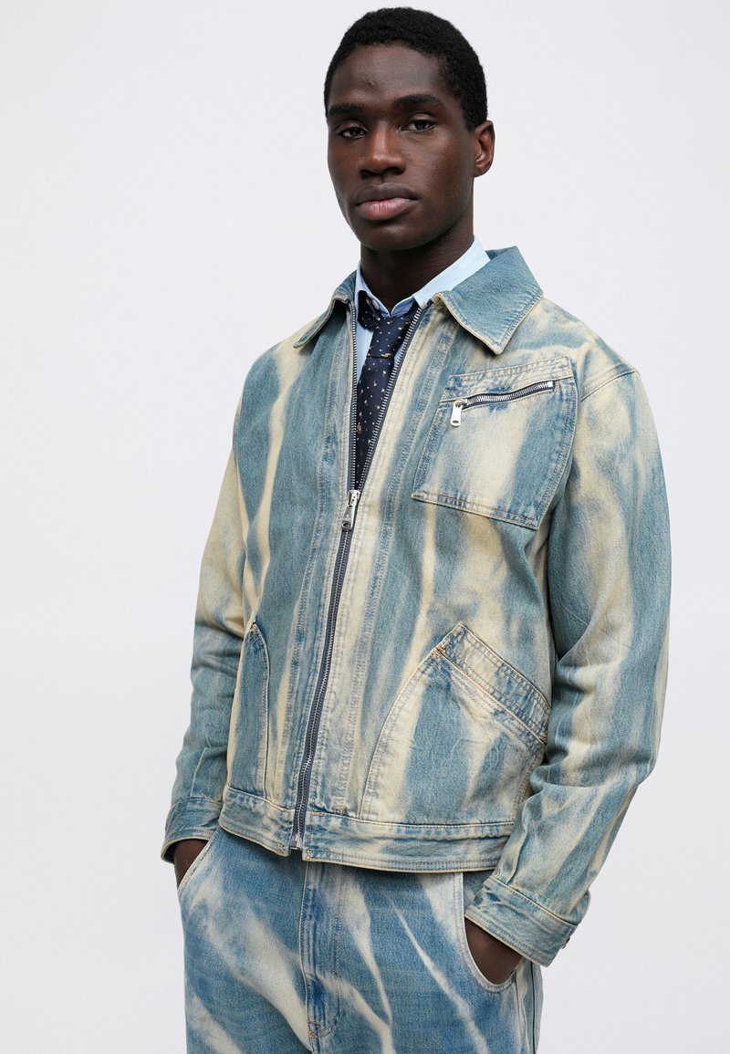 Giubbotto di jeans con motivo a vortice blu chiaro e crema, dotato di zip frontale, due tasche laterali e una tasca sul petto con chiusura a zip.