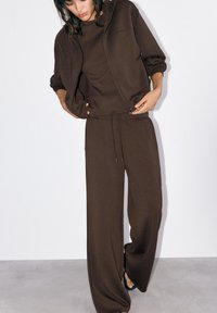 Žena u tamno smeđoj jakni s kapuljačom na patentni zatvarač, u topu dugih rukava u istoj boji i širokim hlačama, stoji naslonjena na jednobojnu bijelu pozadinu.