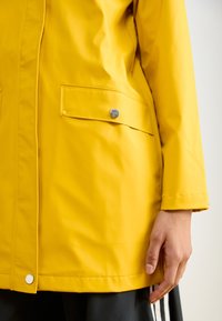 Chaqueta impermeable amarilla con una textura suave, que cuenta con un bolsillo de botón a presión y un acento en tres colores en la manga.
