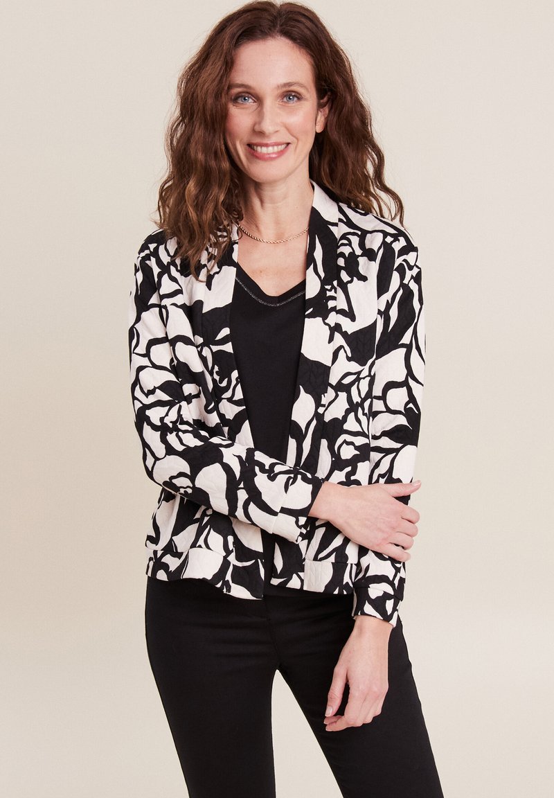 Schwarz-weiß gemusterte Blazerjacke mit floralem Design, kragenlosem Ausschnitt und lässiger Passform, über einem schwarzen Top getragen.