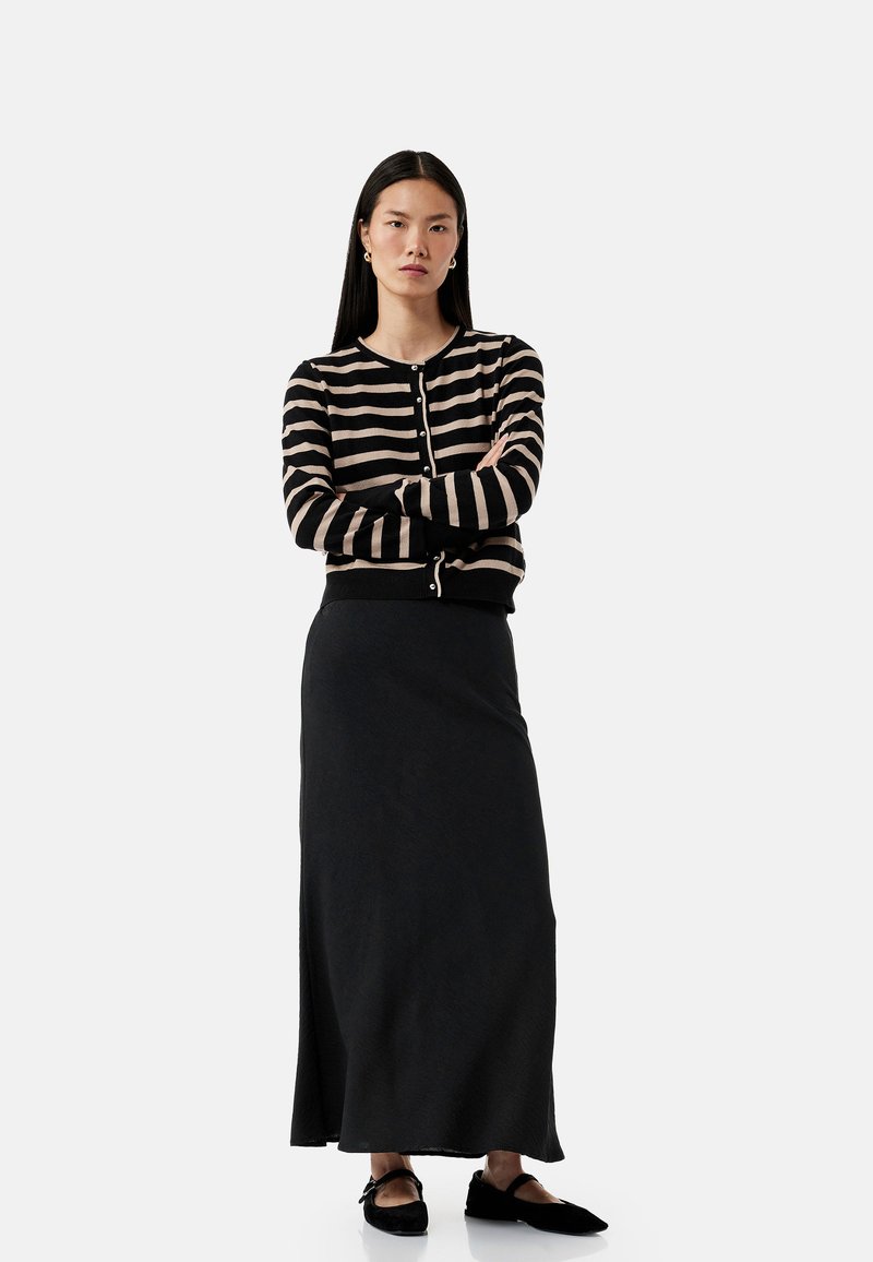 Gestreepte cardigan in zwart en beige, met een sluiting aan de voorkant, gecombineerd met een effen zwarte lange rok en zwarte platte schoenen.