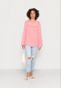 Blusa de manga longa rosa com decote bordado, combinada com jeans com efeito desgastado azul claro. Sandálias brancas rasas e uma bolsa branca texturizada.