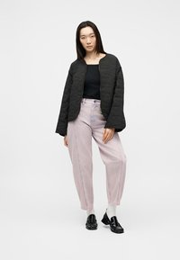 Γυναίκα που φορά μαύρο καπιτονέ μπουφάν, μαύρο τοπ, ανοιχτό ροζ φαρδιά παντελόνια, λευκές κάλτσες και γυαλιστερά μαύρα μοκασίνια, στέκεται μπροστά σε λευκό φόντο.