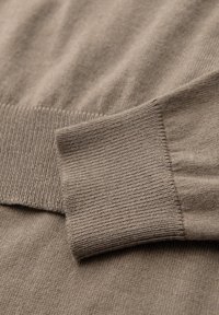 Läheltä kuvattua beigeä villapaitaa, jossa on pehmeä neulepinta, ribbireunus ja sileä pinta. Hienovaraisia väri- ja kuviovaihteluita.