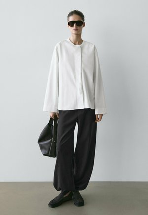 Kobieta w oversize'owej białej koszuli z długim rękawem, czarnych szerokich spodniach, czarnych okularach przeciwsłonecznych, trzymająca czarną torebkę, stojąca na tle jednolitej szarej ściany.