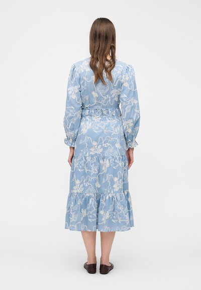 Femme aux cheveux longs bruns portant une robe midi bleue avec une broderie florale blanche et des chaussures plates marron, debout de dos sur un fond uni.
