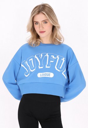 Ung kvinde med blondt hår iført en blå cropped sweatshirt med teksten "JOYFUL LEAGUE" og sorte bukser mod en hvid baggrund.