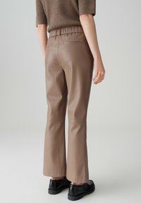 Bruine nep leren wijde broek met een elastische tailleband, voorzien van twee achterzakken en een gladde, matte textuur. Zwarte chunky schoenen worden gedragen.