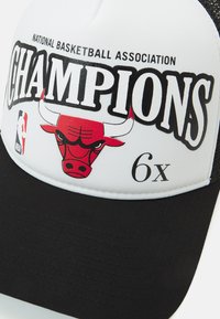 Zwarte en witte pet met een gebogen klep, voorzien van een rood stierenlogo en de tekst "CHAMPIONS". De textuur omvat een gladde stof en mesh-details.