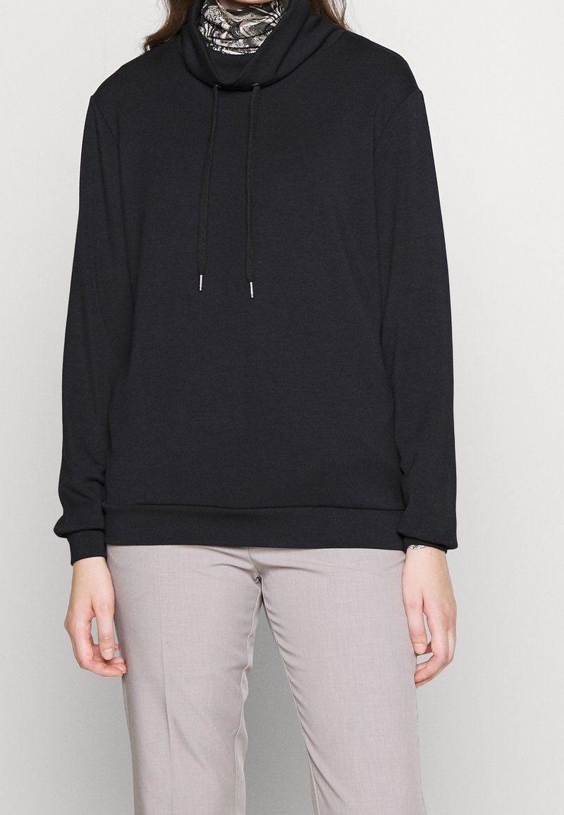 Personne portant un sweat à capuche noir à manches longues avec cordons et un pantalon gris clair, debout devant un fond clair uni.