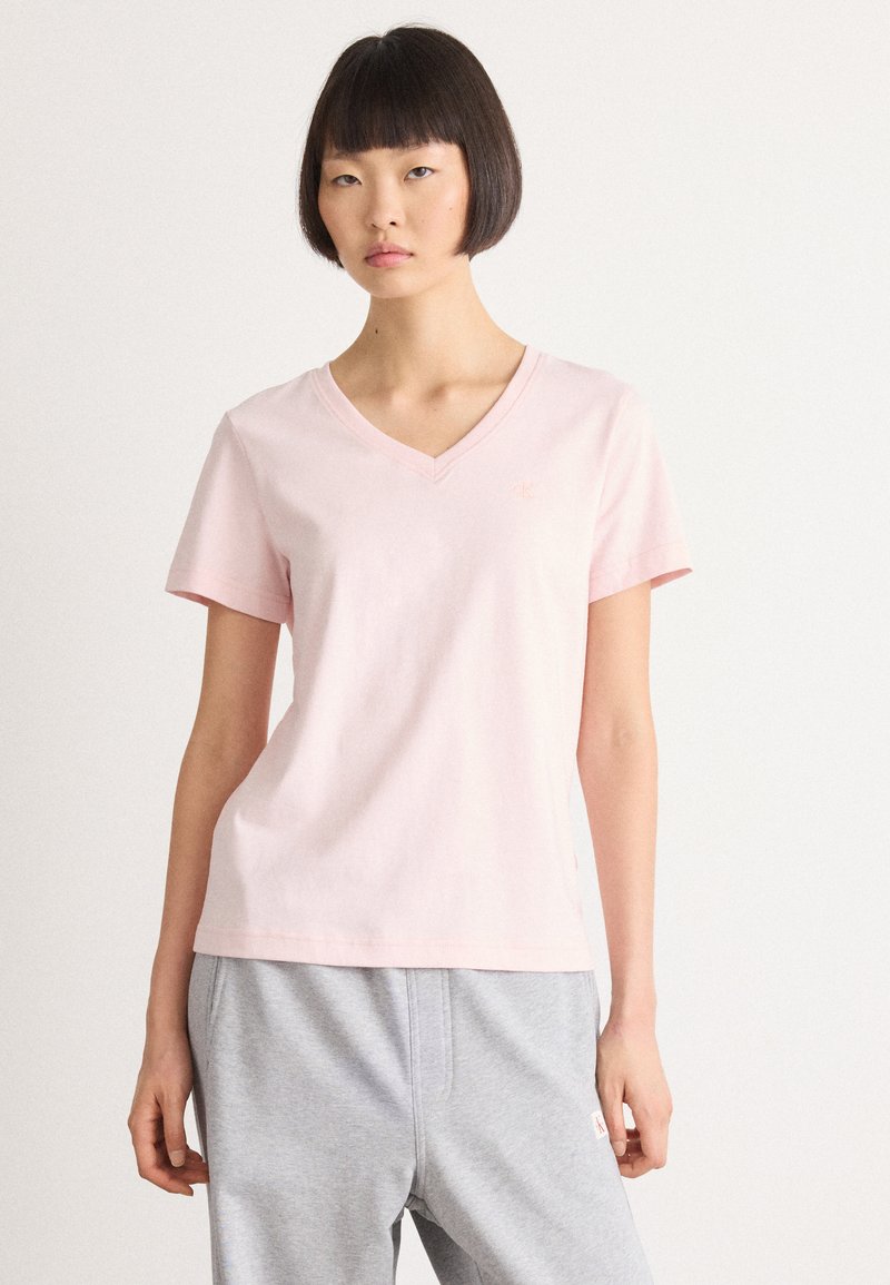 Ung kvinde med kort pagehår iført en lys pink V-hals T-shirt og lyse grå bukser, der står foran en ensfarvet hvid baggrund.