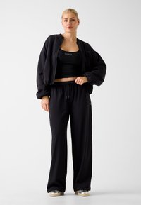 Veste bomber noire raccourcie avec manches surdimensionnées, crop top noir et pantalon noir ample, tous fabriqués en matière douce et légère.