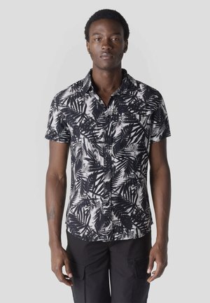 Joven de pie mirando al frente, vistiendo una camisa de manga corta abotonada con un patrón de hojas en blanco y negro y pantalones negros.