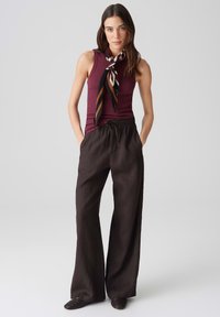 Bordeaux farbenes ärmelloses Oberteil, kombiniert mit lockeren, schwarzen weit geschnittenen Hosen; gemusterter Schal mit Streifen um den Hals; der Stoff wirkt weich und fließend.