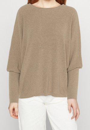 Vrouw in een losse, lange taupe trui met ballonmouwen en een witte broek, staand tegen een effen achtergrond.