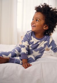 Lilla fleece-nattøj med hvide stjernemønstre, lange ærmer og manchetter. Barnet sidder på en hvid seng med blødt sengetøj.